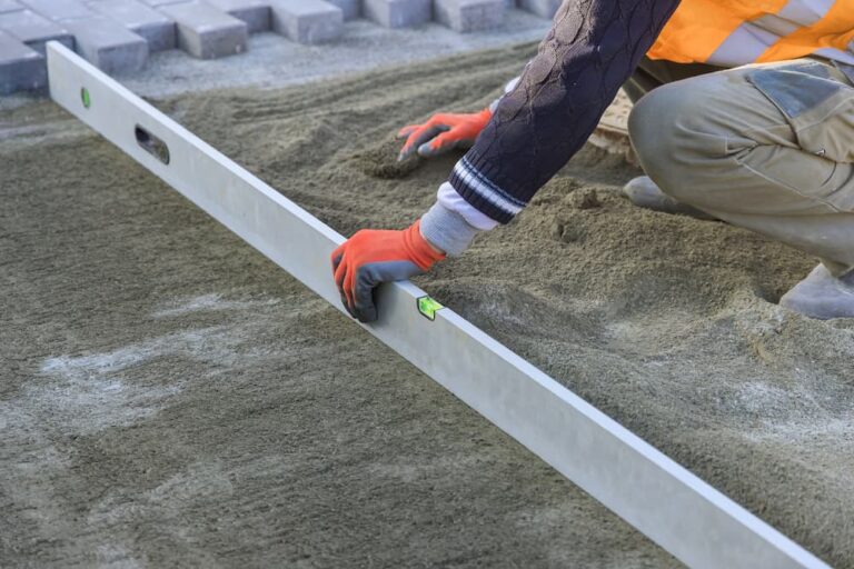 Hardscape contractor using a long spirit level to screed and level a sand base for brick paver installation.